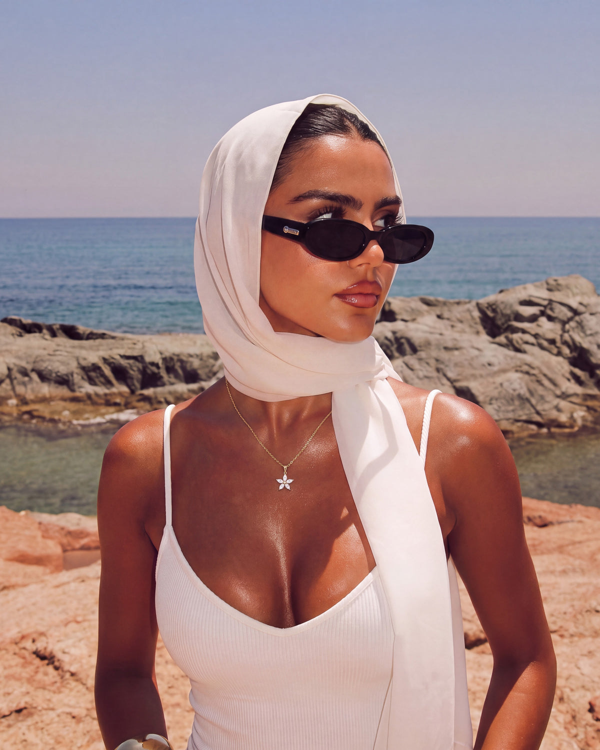 Model in a white headscarf and sunglasses by the sea wearing the Fleur Necklace with flower pendant