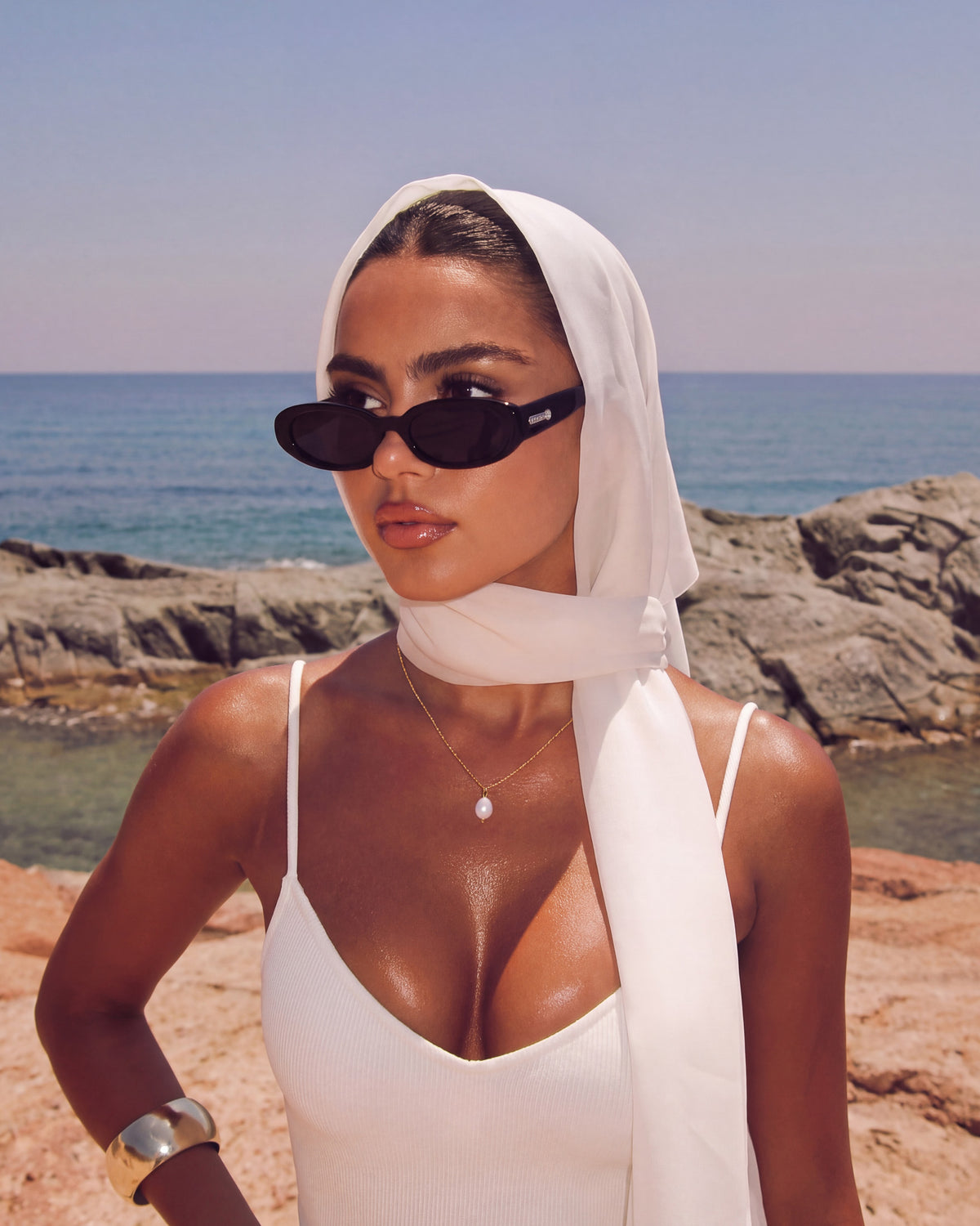 Modelo con pañuelo blanco en la cabeza y lentes de sol junto al mar con el collar Pearl Drop y un top blanco
